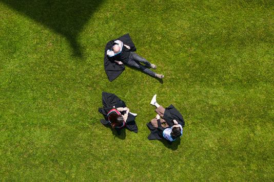 Three QUB graduates with ceremonial gowns on, sitting on the grass taken from directly above